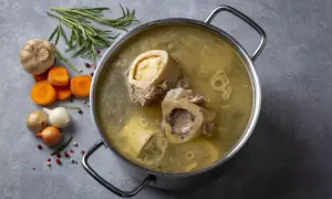 Garnek z bulionem, w którym pływają kości szpikowe na rosół. Obok marchewka, czosnek, cebula i rozmaryn.