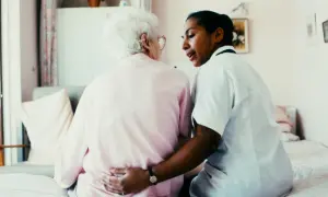 En sjuksköterska, kanske Charlotte Wåhlin, tröstar en äldre kvinna i ett rum.
