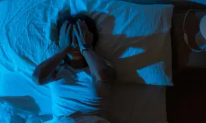 En person ligger i sängen, med händerna över ansiktet, i ett mörkt rum upplyst av blått ljus. En skugga av en hand syns på väggen, som om personen försöker vara fri från tvång.