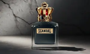 Elegancka butelka perfum Scandal męskie z koroną na nakrętce, stojąca na tle pękniętej ściany.