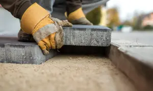 Mani guantate posano un blocco di cemento su un letto di sabbia, creando un nuovo marciapiede in pietra.
