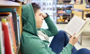Chłopak w kapturze czyta książkę, szukając streszczenia makbeta. Siedzi na podłodze w bibliotece, opierając się o regał.