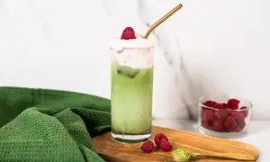 Orzeźwiające matcha z malinami, ozdobione pianką i świeżymi malinami, z dodatkiem proszku matcha na desce.