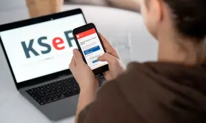 Osoba korzysta z telefonu, by dowiedzieć się, jak wystawić fakturę w KSeF. Na ekranie laptopa widnieje logo KSeF.