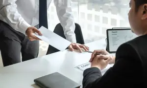 Przełożony wręcza kopertę pracownikowi, który siedzi przy biurku. Na stole leży dokument "CONTRACT" i tablet z raportem.