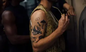 Mężczyzna z tatuażem białego ptaka na czarnym tle. Wzory na ramieniu to m.in. chmurka, parasolka, kość i róża.