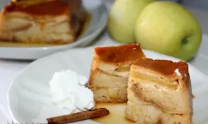 Delicioso pudin de manzana con caramelo, crema batida y canela. Dos porciones servidas en un plato blanco, con dos manzanas verdes al fondo.