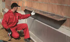 Pracownik w czerwonym kombinezonie montuje okapnik na ścianie zewnętrznej, przygotowując ją do montażu wełny mineralnej.