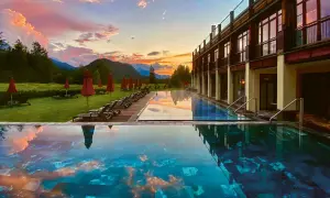 Abendrot über den Alpen spiegelt sich im Infinity-Pool eines der besonderen Hotels Bayern. Liegestühle und rote Sonnenschirme säumen den Rasen.