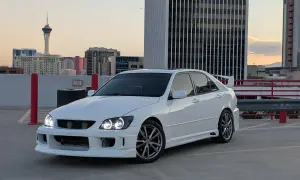 Biała Toyota Altezza z tuningowym body kitem i spojlerem na dachu parkingu, w tle wieżowce i wieża widokowa.
