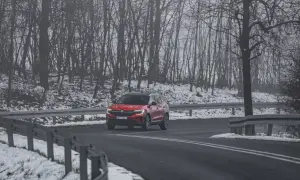 Czerwony SUV jedzie po mokrej drodze, otoczonej ośnieżonym lasem. Kierowca uważa, bo to może być gololedz.