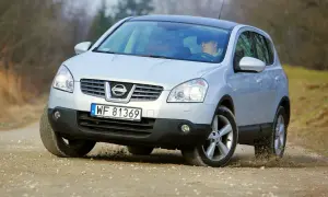Srebrny Nissan Qashqai 1. pokonuje zakręt na piaszczystej drodze, wzbijając kurz.