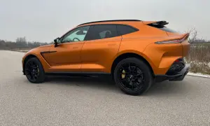 Pomarańczowy Aston Martin DBX z czarnymi felgami i przyciemnianymi szybami na tle pochmurnego nieba.