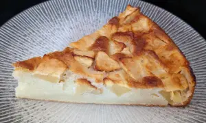 Tranche de clafoutis aux pommes dorée et moelleuse, garnie de morceaux de fruits fondants, sur une assiette texturée.