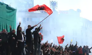 Människor viftar med albanska flaggor och gör fredstecken. En del av folkmassan verkar fira, vilket ger en känsla av stolthet och gemenskap. Detta kan vara en del av fakta om Albanien.