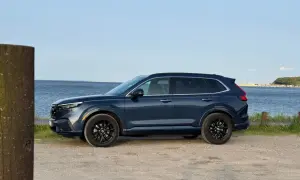Ciemnoniebieski Honda CR-V Hybrid zaparkowany przy wodzie, z widokiem na spokojne morze i łodzie w oddali.
