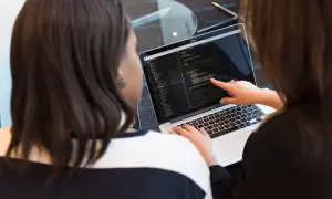 Dwie kobiety analizują kod na laptopie. Jedna wskazuje palcem na ekran, druga pisze. Tak wygląda praca programisty.