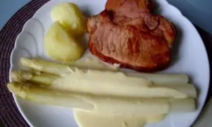 Ein Teller mit gebratenem Schinken, Kartoffeln und Spargel in Sauce Hollandaise. Der Spargel ist in Alufolie gewickelt.