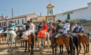 Gente vestida de flamenco a caballo, en un **camino rocío** bajo un cielo azul. Edificios blancos y un campanario al fondo.