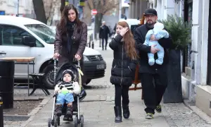 Rodzina spaceruje po mieście, mama pcha wózek z niemowlakiem, a tata niesie drugie dziecko. Wszyscy liroy dzieci wyglądają na szczęśliwych.