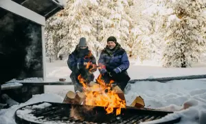 Un couple profite d'un feu de camp dans un paysage de Laponie hivernale, entouré d'arbres enneigés.