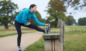Donna in abbigliamento sportivo blu esegue un riscaldamento pre corsa, allungando la gamba su una panchina in un parco.