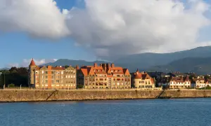 Edificios históricos con tejados rojos bordean el paseo de las grandes villas Getxo, con montañas al fondo y el mar en primer plano.