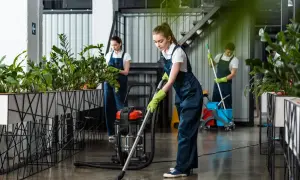 Dziewczyna w granatowym stroju do sprzątania odkurza podłogę. W tle inni pracownicy dbają o czystość i zieleń.