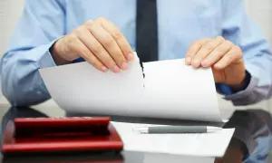 Uomo in camicia blu strappa un documento, simbolo di annullamento proposta di acquisto immobile. Calcolatrice e penna sul tavolo.