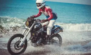 Motocyklista w kasku i kolorowej bluzie śmiga na Honda FMX 650 po plaży, wzbijając fontanny wody.