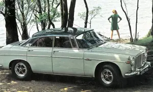 En ljusgrå Rover P5 står parkerad vid en sjö. En kvinna i grön klänning står vid vattenbrynet.