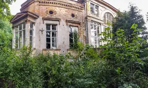 Zarośnięty dworek zakoniczyn z ceglanymi fragmentami i dużymi oknami, otoczony bujną zielenią.