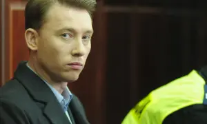 Mężczyzna w ciemnym płaszczu i niebieskiej koszuli patrzy w stronę obiektywu.