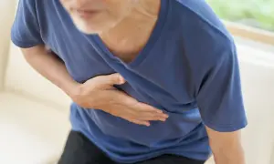 Un homme âgé se tient la poitrine, le visage marqué par la douleur. Sa main droite serre son cœur, comme s'il ressentait une douleur vive, une côte félée peut-être.