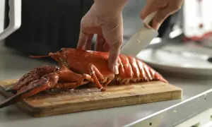 Eine Hand schneidet einen gekochten Hummer auf einem Holzbrett. Die Frage "Hummer kochen wie lange?" wird hier visuell beantwortet.