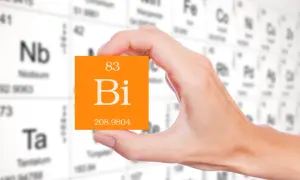 Dłoń trzyma pomarańczową kartkę z symbolem pierwiastka bizmut (Bi) i jego liczbą atomową 83. W tle widać tablicę Mendelejewa.