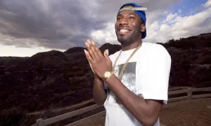 Dougie F. smiles and claps his hands, wearing a blue cap and gold chains, with a dramatic cloudy sky and hills behind him.