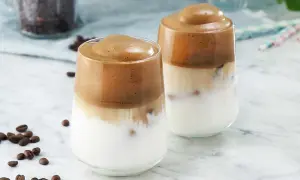 Två glas med krämigt dalgona kaffe, med mjölk och is, toppat med vispad kaffemousse. Kaffebönor sprider sig på marmorbordet.