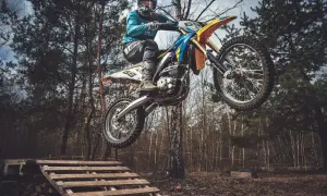 Motocyklista w kasku i goglach skacze na motocyklu enduro nad drewnianą rampą w lesie.
