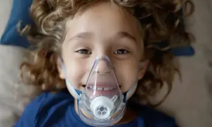 Uśmiechnięte dziecko z kręconymi włosami w masce inhalacyjnej.