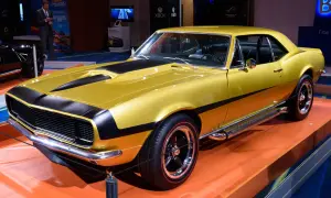 En glänsande gul Chevrolet Camaro SS 1969 med svarta ränder på huven och sidan. Bilen står på en orange yta.