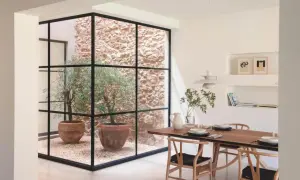 Un comedor moderno con una mesa de madera y sillas de diseño, junto a un patio de luces con olivos en macetas y paredes de piedra.