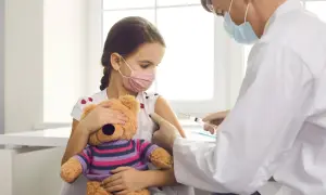 Dziewczynka z maską na twarzy i pluszowym misiem przytula się do ramienia, podczas gdy lekarz wykonuje szczepienia covid.