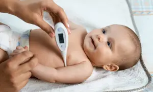 Mama mierzy temperaturę maluszkowi. Cyfrowy termometr pokazuje 36.6°C, co jest prawidłową temperaturą. Dziecko spokojnie leży na białym ręczniku.