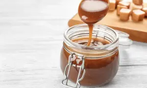 Une cuillère verse un coulis de caramel onctueux dans un bocal en verre. Des bonbons au caramel sont disposés sur une planche en bois en arrière-plan.