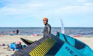 Mężczyzna w kasku i piance na plaży, leżące latawce do kitesurfingu, morze i inni sportowcy.