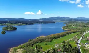Weite Sicht auf den Lipno Stausee mit bewaldeten Hügeln, grünen Wiesen und kleinen Häusern am Ufer.