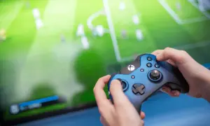 Ręce trzymające niebieski kontroler Xbox, grające w grę piłkarską. Ekran telewizora pokazuje zielone boisko z zawodnikami, tworząc efektowną teselację.