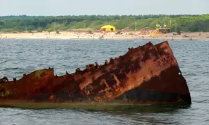 Zardzewiała rufa statku wystaje z wody na tle plaży pełnej ludzi i zielonego lasu.