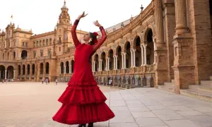 Bailarina con vestido rojo voluminoso, brazos en alto, ejecutando sevillanas lentas frente a un edificio histórico con arcos.